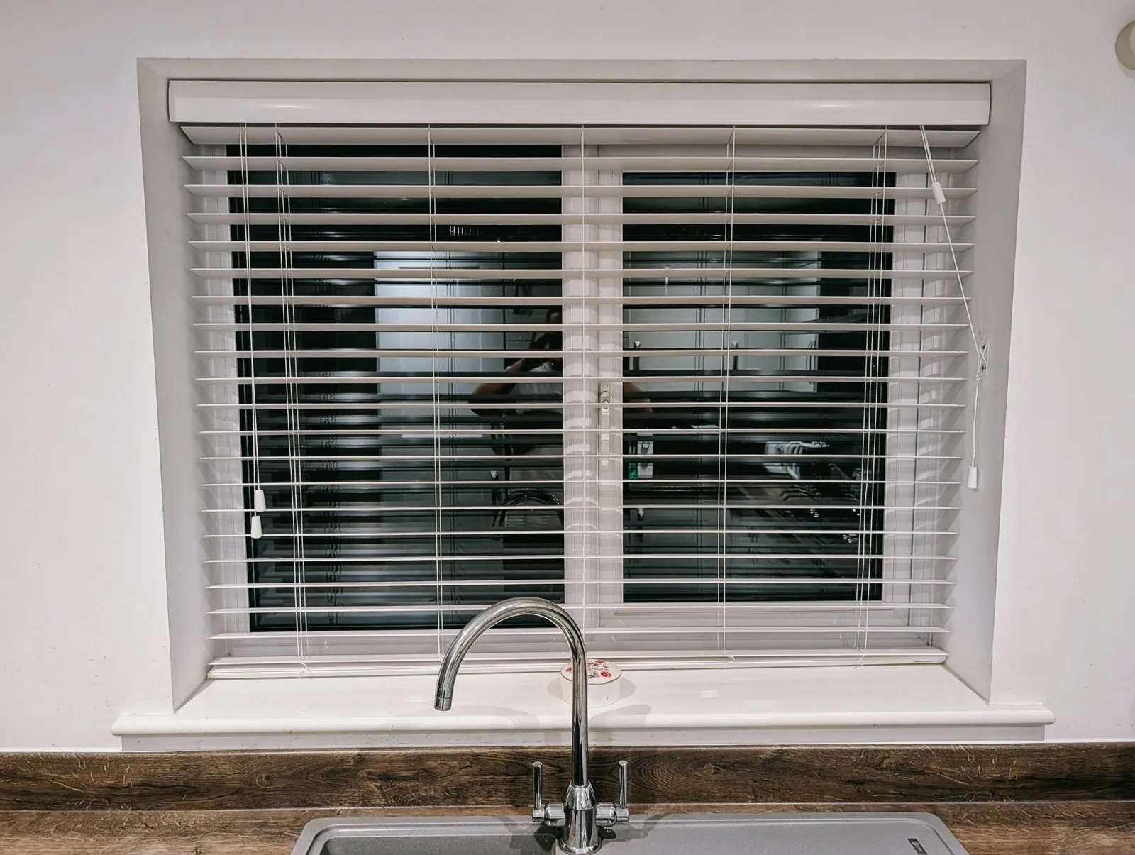 White venetian blind fitted in the recess of a wide kitchen window above a sink